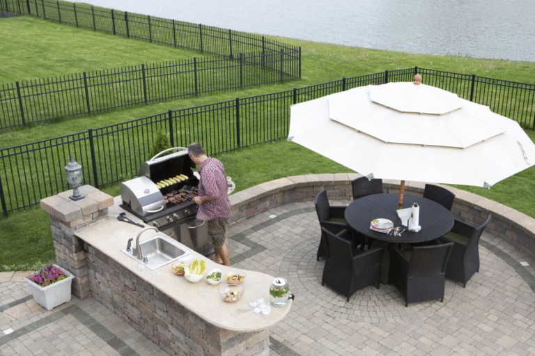 a man grilling out back of his water front property, with an umbrella covered dining area nearby and an aluminum fence surround both his and his neighbors property. They chose to leave space between their fences to allow lawn maintenance and water access