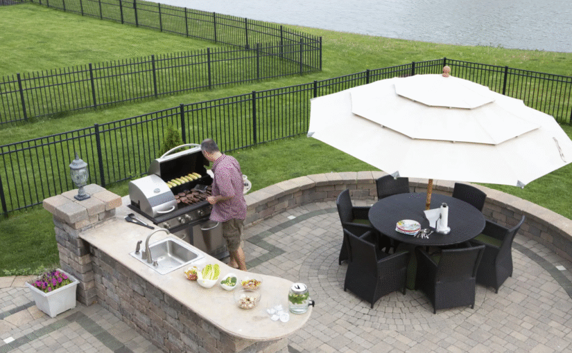 a man grilling out back of his water front property, with an umbrella covered dining area nearby and an aluminum fence surround both his and his neighbors property. They chose to leave space between their fences to allow lawn maintenance and water access