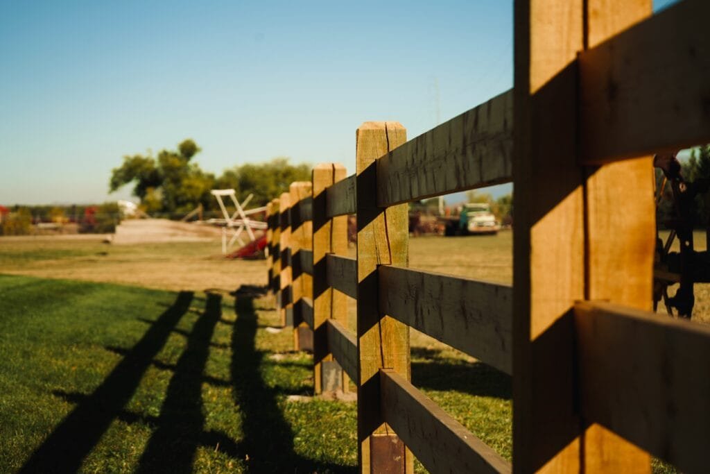 a 3 rail wooden farm fence with a shadow casted on the grass of a farm