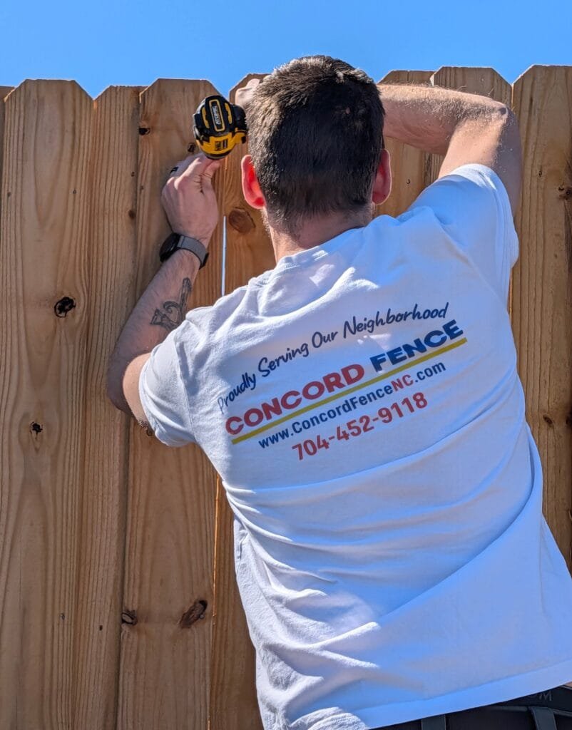 jake from concord fence repairs a board on board privacy fence. The back of his tshirt shows the concord fence logo, stating "proudly serving the neighborhood" with their contact information listed below